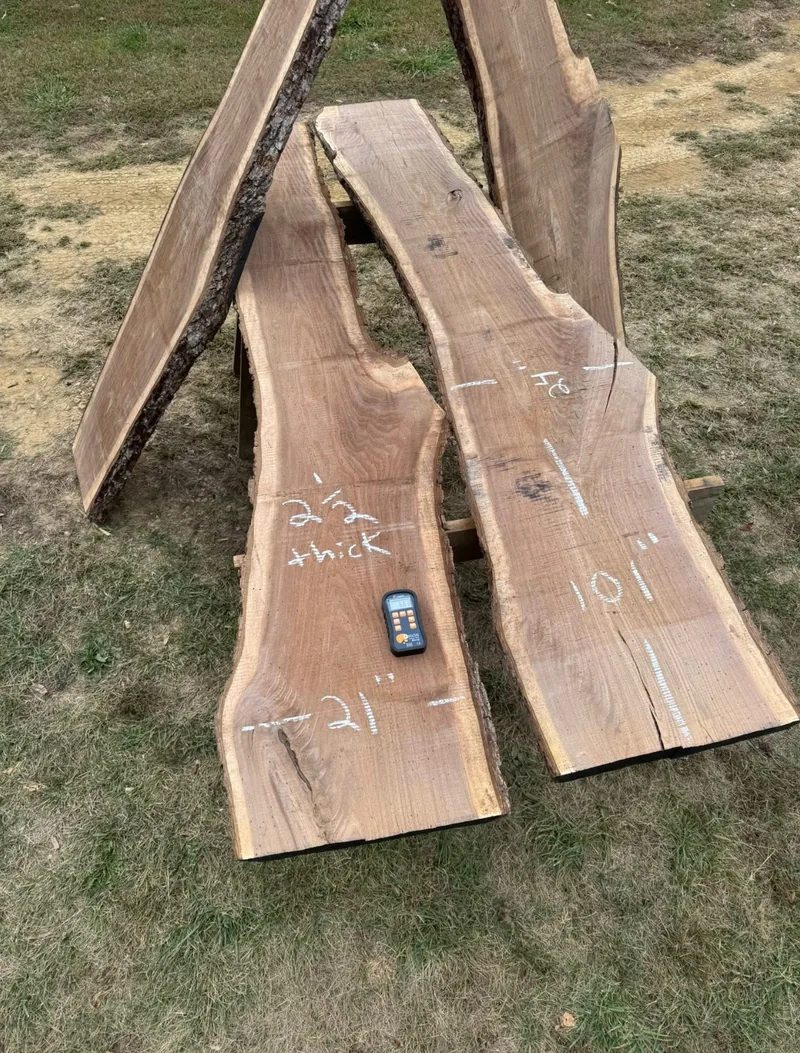 Live-edge hardwood slabs on sawhorses, chalk-marked with width and thickness measurements beside a moisture meter